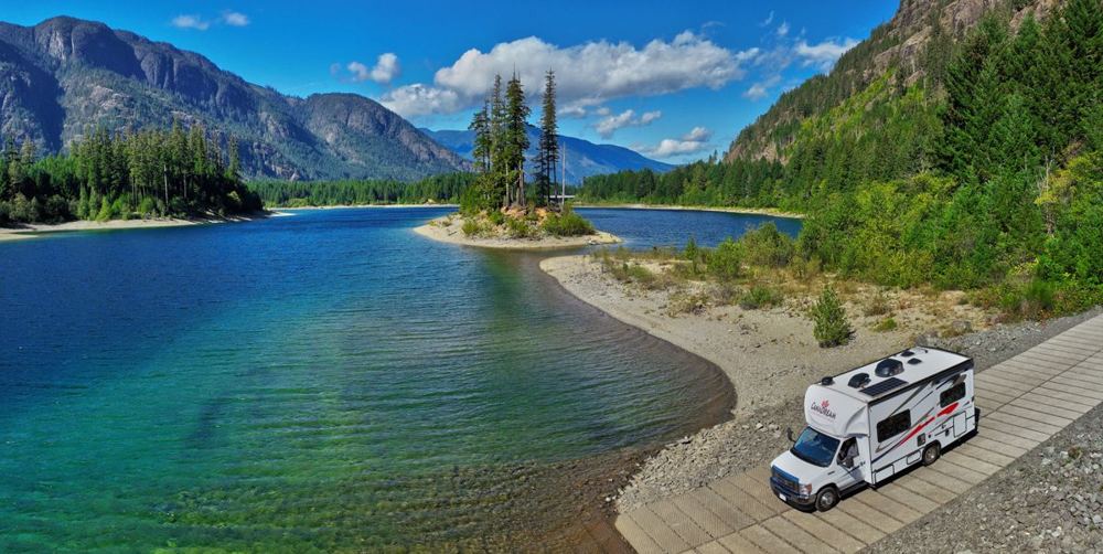 Una autocaravana aparcada a la orilla de un lago de agua verde entre montañas, vista desde el aire