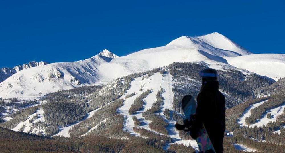 Un snowboarder de espaldas, con la tabla en la mano, mira una montaña nevada surcada de pistas